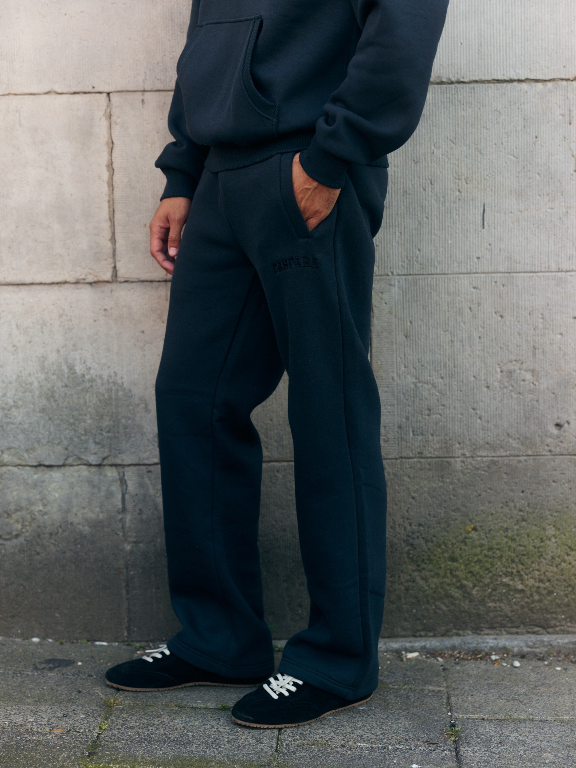 Man wearing black straight-leg sweatpants and a matching hoodie standing against a stone wall.